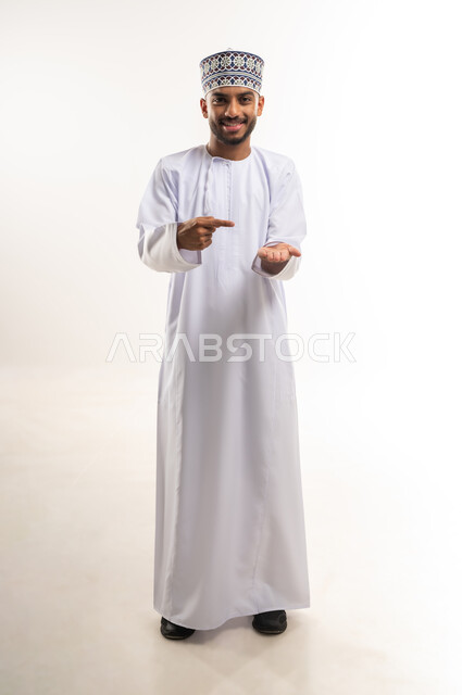 رفع شيء معين, تعابير حمل و إمساك عنصر محدد, النظر إلى الكاميرا بإيماءات السعادة و السرور, الإشارة إلى هدف ما، بورتريه لرجل عربي خليجي عماني يرتدي الدشداشة والكمة يشير بإصبع السبابة إلى يده, الجسم بالطول الكامل، خلفية بيضاء