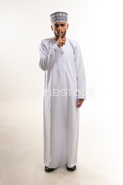 التوقف عن الكلام و الأمر بالصمت و عدم إصدار صوت, النظر إلى الكاميرا بإيماءات الجدية و الحزم، بورتريه لرجل عربي خليجي عماني يرتدي الدشداشة والكمة يرفع إصبع السبابة إلى فمه بتعابير طلب الهدوء, الجسم بالطول الكامل, خلفية بيضاء