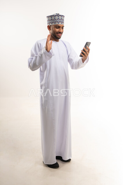 التواصل مع العائلة والأصدقاء, مشاركة الأخبار والأحاديث الجيدة, استخدام جهاز تقني حديث ومتطور، بورتريه لرجل عربي خليجي عماني يرتدي الدشداشة والكمة يقوم بإجراء مكالمة فيديو عبر الهاتف المحمول ويرفع يده لإلقاء التحية والسلام, الجسم بالطول الكامل, خلفية بيضاء