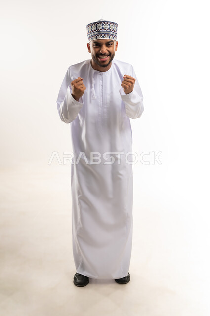 النظر الى الكاميرا بإيماءات السعادة والسرور, تعابير الفوز و تحقيق الأهداف، الفرح لسماع خبر ما, بورتريه لشاب عربي خليجي عماني يرتدي الدشداشة والكمة يرفع يديه بحركات الحماس, الجسم بالطول الكامل, خلفية بيضاء