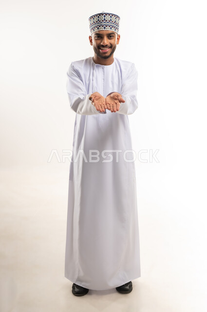 Presentation concept, looking at the camera with gestures of happiness and pleasure, holding an item, portrait of an Arab Gulf Omani man wearing a dishdasha and a kummah extending his hands forward with an expression of lifting an object, full body, white background