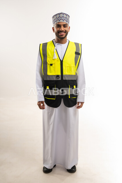 النظر إلى الكاميرا بإيماءات السعادة والسرور، العمل في القطاع الهندسي, الإعمار بسواعد أبناء الوطن, المتابعة والإشراف على المشاريع الهندسية, بورتريه لمهندس عربي خليجي عماني يرتدي الدشداشة والكمة مع سترة الحماية يقف باستقامة واعتدال, الجسم بالطول الكامل, خلفية بيضاء