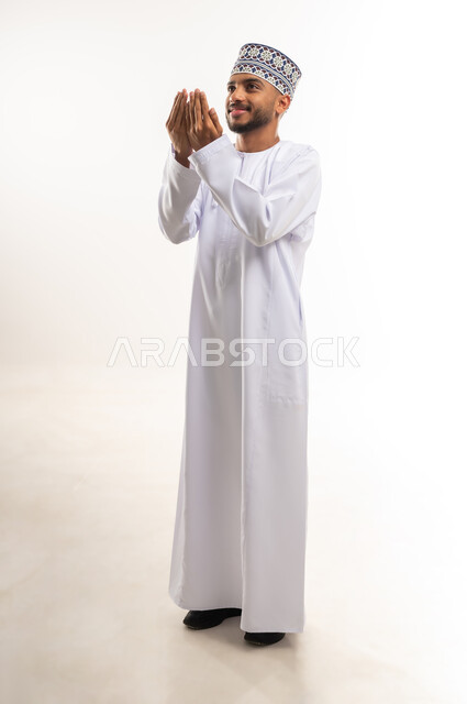 مفهوم العبادة والتقرب إلى الله تعالى, الاجتهاد بالأدعية والطاعات والأذكار, بورتريه لرجل عربي خليجي عماني يرتدي الدشداشة والكمة يرفع يديه للأعلى بتعابير الدعاء, الجسم بالطول الكامل, خلفية بيضاء