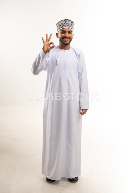 التعبير عن الجودة والاتقان, النظر إلى الكاميرا بإيماءات السعادة والسرور, مفهوم الموافقة والقبول والنصيحة, بورتريه لرجل عربي خليجي عماني يرتدي الدشداشة والكمة يرفع يده بتعابير الامتياز, الجسم بالطول الكامل, خلفية بيضاء