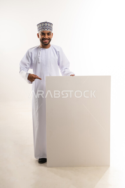 النظر إلى الكاميرا بإيماءات السعاة والسرور, موك اب مستطيل أبيض، استخدام لوحات توضيحية وتعليمية، إعلانات وعروض تسويقية, بورتريه لرجل عربي خليجي عماني يرتدي الدشداشة والكمة يمسك بيده لوحة إعلانية فارغة ويشير إليها بإصبع السبابة, الجسم بالطول الكامل, خلفية بيضاء