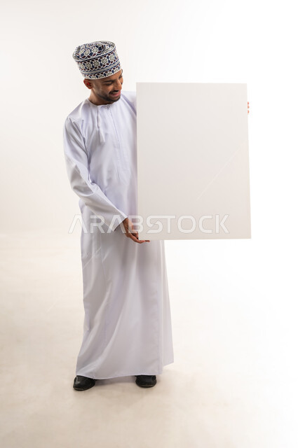 لوح إعلاني فارغ, موك اب مستطيل أبيض، استخدام لوحات توضيحية ووسائل تعليمية، إعلانات وعروض تسويقية, بورتريه لرجل عربي خليجي عماني يرتدي الدشداشة والكمة يمسك بيده لوحة إعلانية فارغة وينظر إليها, الجسم بالطول الكامل, خلفية بيضاء