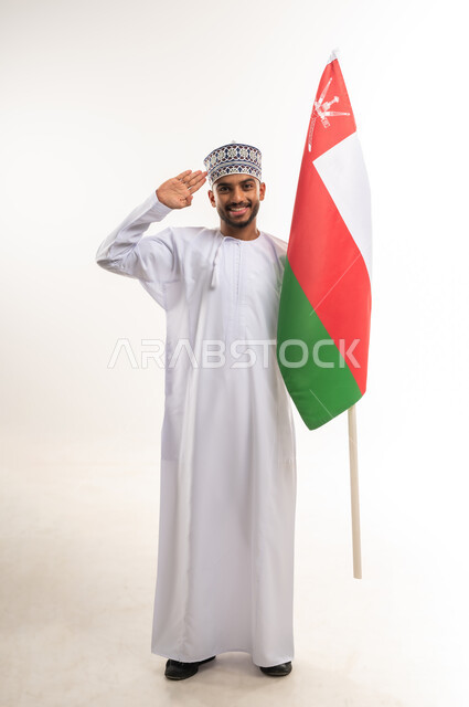 الفخر والاعتزاز بالهوية العمانية, ذكرى اليوم الوطني العماني 18 نوفمبر, الاحتفال بالأعياد والمناسبات الوطنية, بورتريه لرجل عربي خليجي عماني مبتسم يرتدي الدشداشة والكمة يمسك علم سلطنة عمان ويرفع يده إلى رأسه بتعابير إلقاء التحية, الجسم بالطول الكامل, خلفية بيضاء