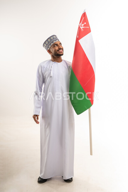 مفهوم الحب والانتماء، الاحتفال بالأعياد والمناسبات الوطنية, الفخر والاعتزاز بالهوية العمانية, استذكار اليوم الوطني العماني 18 نوفمبر, بورتريه لرجل عربي خليجي عماني مبتسم يرتدي الدشداشة والكمة يمسك علم سلطنة عمان وينظر إليه, الجسم بالطول الكامل, خلفية بيضاء