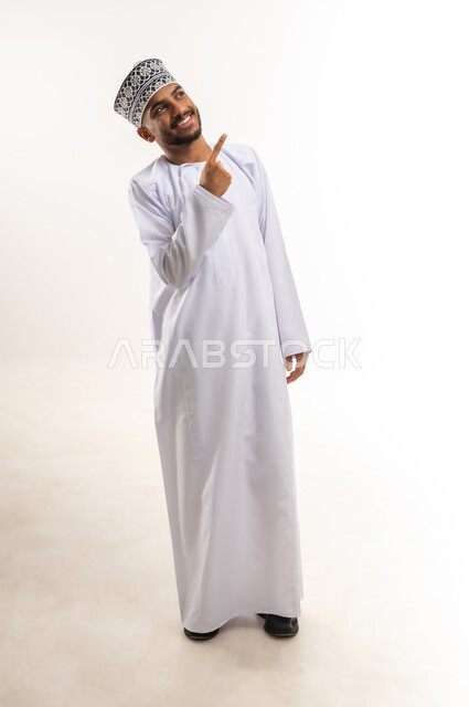 Indication and explanation of a specific place, pointing to a specific location, locating an object, portrait of a smiling Arab Gulf Omani man wearing a dishdasha and a kummah pointing with his index finger to something above, full body, white background