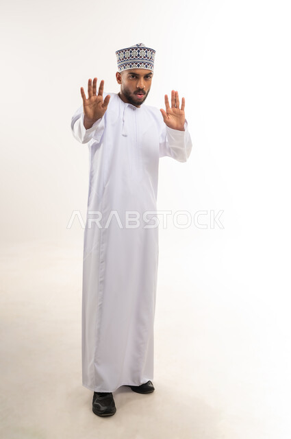 طلب التوقف و الامتناع و عدم الاقتراب, حماية النفس من ضرر خارجي, بورتريه لرجل عربي خليجي عماني يرتدي الدشداشة والكمة ينظر إلى الكاميرا و يرفع يديه بتعابير الخوف و الابتعاد, الجسم بالطول الكامل, خلفية بيضاء