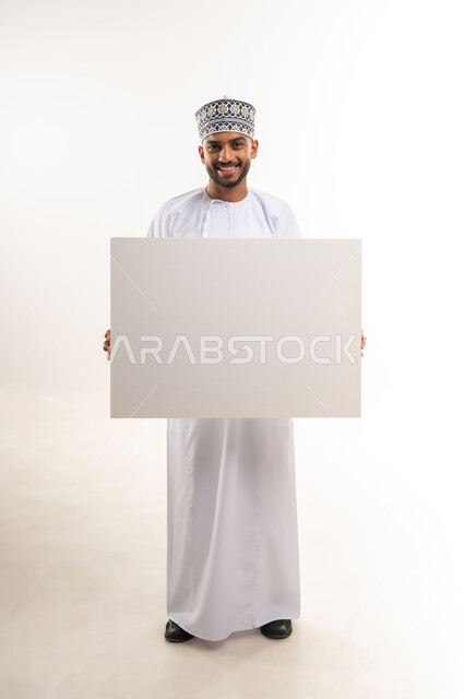 موك اب مستطيل أبيض، لوح إعلاني فارغ، استخدام لوحات توضيحية ووسائل تعليمية، إعلانات وعروض تسويقية, النظر إلى الكاميرا بإيماءات السعادة والسرور, بورتريه لرجل عربي خليجي عماني يرتدي الدشداشة والكمة يمسك بيده لوحة إعلانية فارغة, الجسم بالطول الكامل, خلفية بيضاء