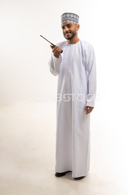 النظر إلى الكاميرا بإيماءات السعادة والسرور، الاستجابة لحالات الطوارئ، المتابعة والإشراف على العمل, استعمال التقنيات الحديثة، بورتريه لرجل عربي خليجي عماني يرتدي الدشداشة والكمة يقوم باستخدام جهاز الإرسال والاستقبال اللاسلكي, الجسم بالطول الكامل، خلفية بيضاء