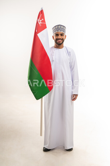 الاحتفال بالأعياد والمناسبات الوطنية, مفهوم الحب والانتماء, الفخر والاعتزاز بالهوية العمانية, استذكار اليوم الوطني العماني 18 نوفمبر, بورتريه لرجل عربي خليجي عماني مبتسم يرتدي الدشداشة والكمة يمسك علم سلطنة عمان, الجسم بالطول الكامل, خلفية بيضاء