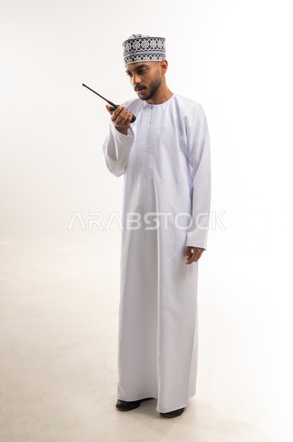 مفهوم إدارة المشاريع عن بعد، الاستجابة لحالات الطوارئ، المتابعة والإشراف على العمل, استخدام التقنيات الحديثة في الأعمال، بورتريه لرجل عربي خليجي عماني يرتدي الدشداشة والكمة يقوم باستخدام جهاز الإرسال والاستقبال اللاسلكي, الجسم بالطول الكامل، خلفية بيضاء
