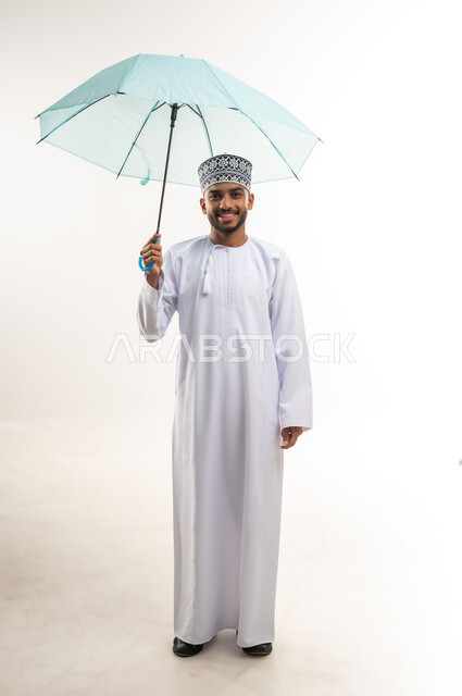 النظر إلى الكاميرا بإيماءات السعادة والسرور, التظلل من أشعة الشمس, الحماية من الأمطار في الشتاء، بورتريه لرجل عربي خليجي عماني مبتسم يرتدي الدشداشة والكمة يحمل بيده مظلة زرقاء, الجسم بالطول الكامل, خلفية بيضاء
