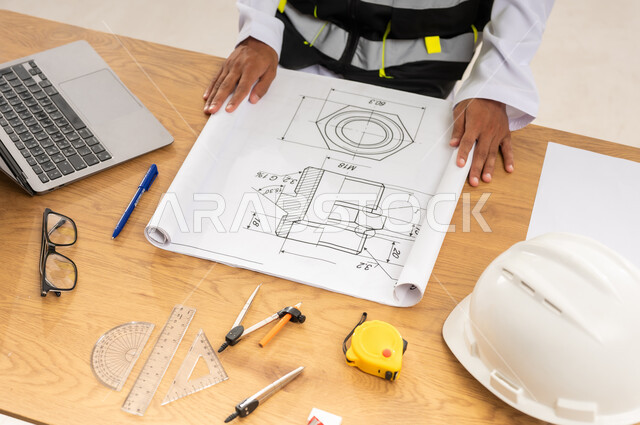 Studying and reviewing construction plans, integrating modern technical devices into engineering works, using engineering tools and equipment, working in the engineering sector, close-up of an Arab Gulf Omani engineer wearing a protective vest placing the architectural plan in front of him