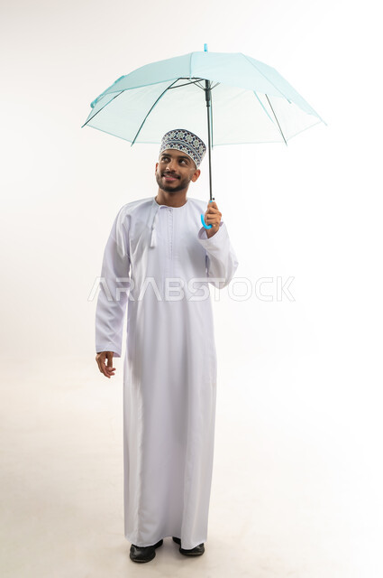 التظلل من أشعة الشمس, الحماية من الأمطار في الشتاء، النظر إلى الأعلى بإيماءات السعادة والسرور, بورتريه لرجل عربي خليجي عماني مبتسم يرتدي الدشداشة والكمة يحمل بيده مظلة زرقاء, الجسم بالطول الكامل, خلفية بيضاء
