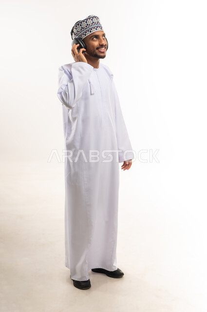 الاندماج مع الألحان الموسيقية, استخدام التقنيات الحديثة للتسلية, قضاء الوقت الممتع, الاستمتاع بسماع الموسيقى المفضلة, بورتريه لشاب عربي خليجي عماني يرتدي الدشداشة والكمة يضع سماعات الرأس الصوتية ينظر إلى شيء ما بالأعلى, الجسم بالطول الكامل, خلفية بيضاء