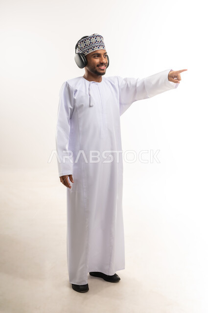 Pointing with index finger at something, using modern and advanced technologies for entertainment and amusement, having a good time, enjoying listening to favorite music, portrait of a young Arab Gulf Omani man wearing a dishdasha and a kummah, wearing audio headphones, full body, white background