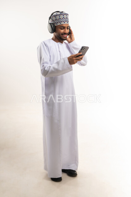 استخدام الأجهزة التقنية الحديثة والمتطورة للتسلية والترفيه, قضاء الوقت الممتع, الاستمتاع بسماع الموسيقى المفضلة, بورتريه لشاب عربي خليجي عماني يرتدي الدشداشة والكمة يضع سماعات الرأس الصوتية ويمسك الهاتف المحمول بيده, الجسم بالطول الكامل, خلفية بيضاء