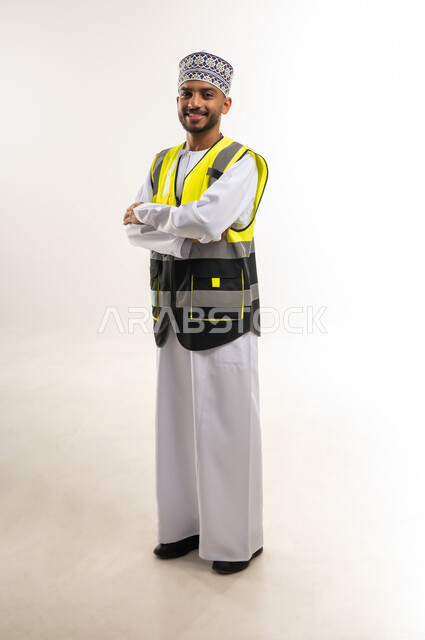 الإعمار بسواعد أبناء الوطن, العمل في القطاع الهندسي, المتابعة والإشراف على المشاريع الهندسية, بورتريه لمهندس عربي خليجي عماني يرتدي الدشدشة والكمة مع سترة الحماية ينظر إلى الكاميرا بإيماءات السعادة وأيدي متقاطعة, الجسم بالطول الكامل, خلفية بيضاء