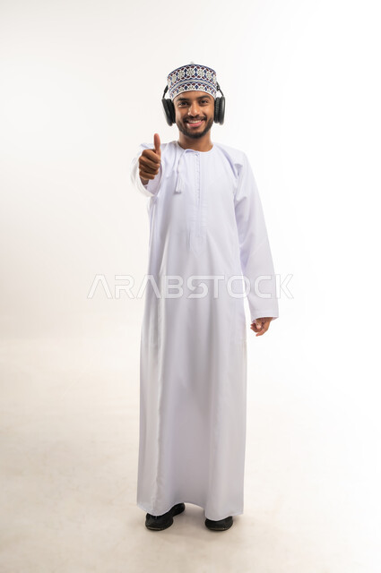 تعابير الجودة والامتياز, استخدام التقنيات الحديثة للتسلية والترفيه, النظر إلى الكاميرا بإيماءات السعادة والسرور, بورتريه لشاب عربي خليجي عماني يرتدي الدشداشة والكمة يضع سماعات الرأس الصوتية ويشير بإصبع الإبهام للأعلى, الجسم بالطول الكامل, خلفية بيضاء