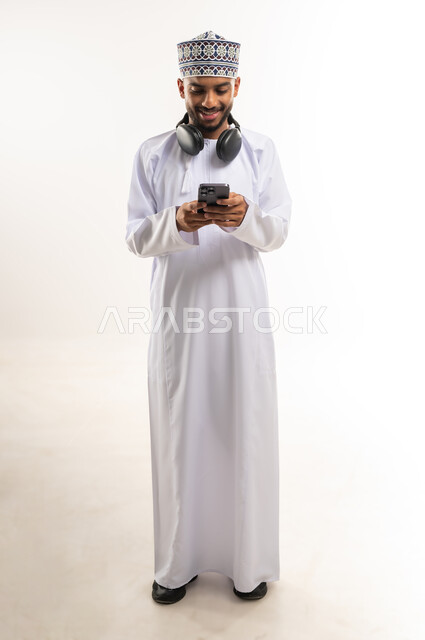 Having a good time, using modern and advanced technical devices for entertainment and amusement, enjoying listening to favorite music, portrait of a young Arab Gulf Omani man wearing a dishdasha and a kummah, putting on audio headphones and holding a mobile phone in his hand, full body, white background