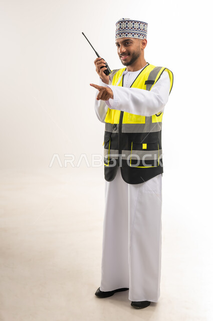 Pointing index finger at something, managing and supervising construction projects, working in engineering sector, portrait of an Arab Gulf Omani engineer wearing a dishdasha and a kimono with a protective vest using a walkie-talkie, full body, white background