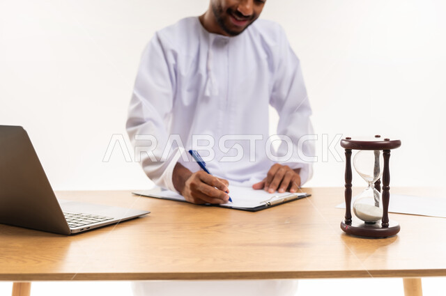 مفهوم إدارة الوقت, إعداد التقارير الخاصة بالعمل, دمج التقنيات الحديثة في الأعمال المكتبية, إنجاز المهام في الوقت المحدد, بورتريه مقرب لرجل عربي خليجي عماني يرتدي الدشداشة يضع أمامه ساعة رملية ويقوم بكتابة وتدوين الملاحظات وبجانبه الحاسوب المحمول, خلفية بيضاء