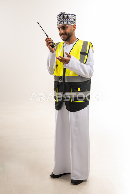 التواصل مع فريق العمل عن بعد, الإدارة والإشراف على المشاريع الإنشائية, العمل في القطاع الهندسي, بورتريه لمهندس عربي خليجي عماني يرتدي الدشداشة والكمة مع سترة الحماية يقوم باستخدام جهاز الاتصال اللاسلكي, الجسم بالطول الكامل, خلفية بيضاء