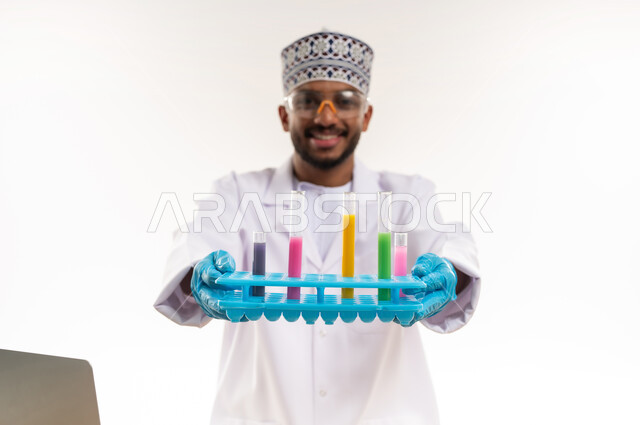 النظر إلى الكاميرا بإيماءات السعادة والسرور, مفهوم الكيمياء الحيوية، دمج الأجهزة الحديثة والتكنولوجيا في الحياة العملية, القيام بالتجارب العلمية, بورتريه مقرب لأخصائي مختبر عربي خليجي عماني يرتدي المعطف الأبيض والكمة يمسك بيديه أنابيب الاختبار, خلفية بيضاء