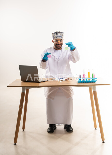 دمج الأجهزة الحديثة والتكنولوجيا في الحياة العملية, مفهوم الكيمياء الحيوية، القيام بالتجارب والبحوث العلمية, بورتريه لأخصائي مختبر عربي خليجي عماني يرتدي المعطف الأبيض والكمة يقوم باستعمال أنابيب الاختبار, خلفية بيضاء