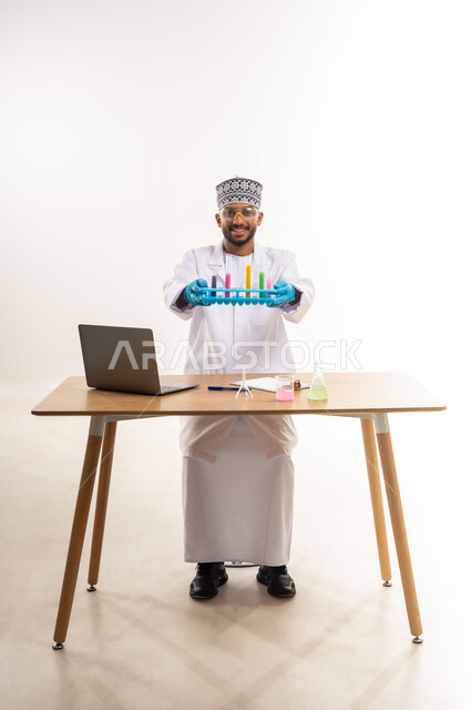 القيام بالتجارب والبحوث العلمية, دمج الأجهزة الحديثة والتكنولوجيا في الحياة العملية, مفهوم الكيمياء الحيوية, بورتريه لأخصائي مختبر عربي خليجي عماني يرتدي المعطف الأبيض والكمة يمسك بيديه أنابيب الاختبار, خلفية بيضاء