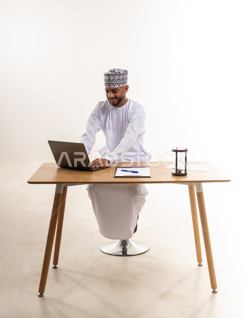 إنجاز المهام بدقة في الزمن المحدد, دمج التقنيات الحديثة في الأعمال المكتبية, مفهوم إدارة الوقت والجدول الزمني, بورتريه لرجل عربي خليجي عماني يرتدي الدشداشة والكمة يضع أمامه ساعة رملية ويقوم بالعمل على جهاز الحاسوب المحمول, خلفية بيضاء