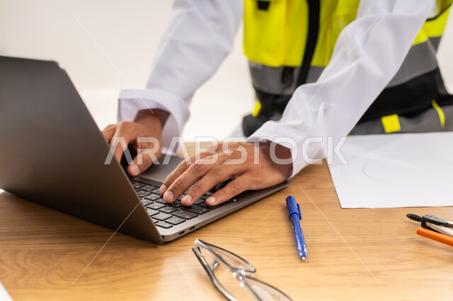 المتابعة والإشراف على المشاريع الإنشائية، تطور و نمو القطاع الهندسي, دمج التقنيات والتكنولوجيا الحديثة بالأعمال الهندسية, بورتريه مقرب لمهندس عربي خليجي عماني يرتدي سترة الحماية يقوم بالكتابة على جهاز الحاسوب المحمول, خلفية بيضاء