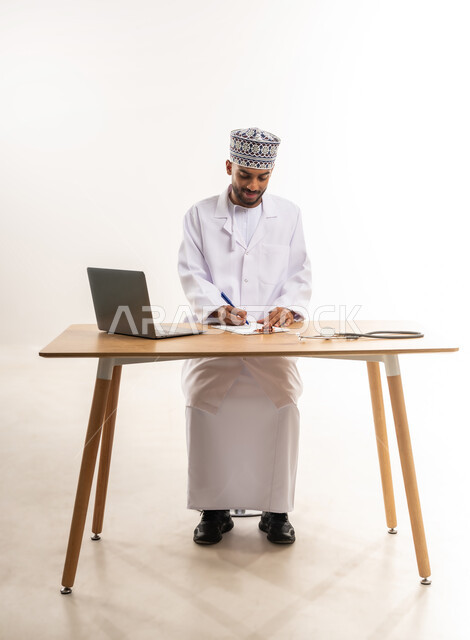 العمل في القطاع الطبي, تقديم خدمات الرعاية الصحية للمرضى, إجراء الاستشارات عن بعد, دمج الأجهزة التقنية الحديثة بالطب, بورتريه لطبيب عربي خليجي عماني يرتدي المعطف الأبيض والكمة يقوم بكتابة وصفة طبية, خلفية بيضاء