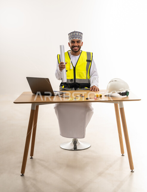 النظر إلى الكاميرا بإيماءات السعادة والسرور, متابعة المخططات الإنشائية, دمج الأجهزة التقنية الحديثة بالأعمال الهندسية, بورتريه لمهندس عربي خليجي عماني يرتدي الدشداشة والكمة مع سترة الحماية يجلس على الطاولة ويمسك بيده المخطط المعماري, خلفية بيضاء