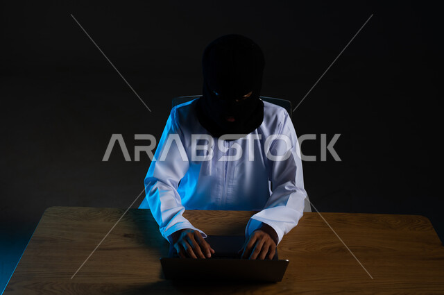 استخدم جهاز الحاسوب لأغراض غير مشروعة, قرصنة وسرقة البيانات, تدمير الحسابات الرقمية، مفهوم الجرائم الالكترونية, القدرة على تهكير الأجهزة, بورتريه مقرب لهاكر عربي خليجي عماني ملثم يرتدي الدشداشة يعمل في مجال الهكر الغير قانوني، خلفية سوداء