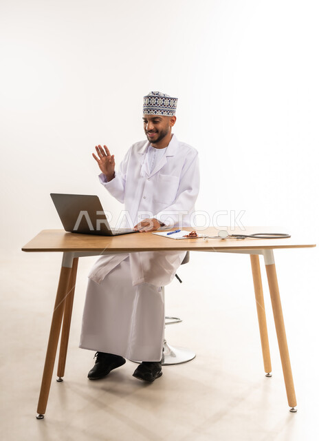 إجراء الاستشارات عن بعد، تقديم خدمات الرعاية الصحية للمرضى, دمج الأجهزة التقنية الحديثة بالطب, بورتريه لطبيب عربي خليجي عماني يرتدي المعطف الأبيض والكمة يقوم بإجراء مكالمة فيديو عبر الحاسوب المحمول ويرفع يده بتعابير السلام, خلفية بيضاء