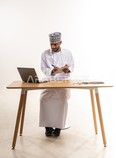 تقديم خدمات الرعاية الصحية للمرضى, إجراء الاستشارات عن بعد، العمل في القطاع الطبي, دمج الأجهزة التقنية الحديثة بالطب, بورتريه لطبيب عربي خليجي عماني يرتدي المعطف الأبيض والكمة ينظر إلى الحاسوب المحمول  ويقوم بكتابة وصفة طبية, خلفية بيضاء