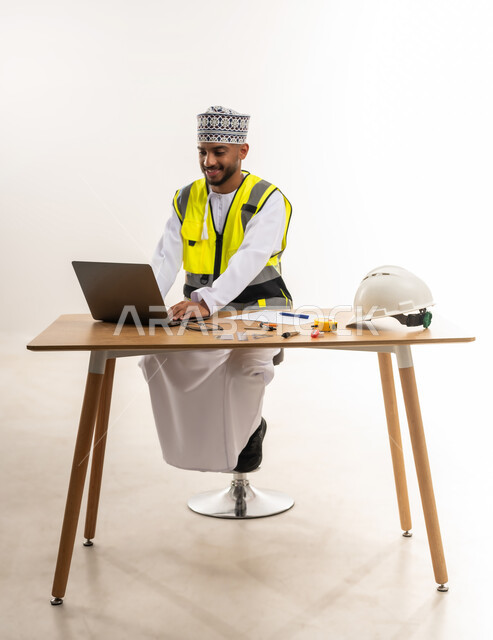 Integrating modern technical devices with engineering work, working in the engineering sector, following up on projects and construction plans, portrait of an Arab Gulf Omani engineer wearing a dishdasha and a cloak with a protective jacket sitting at the table and working on a laptop, white background