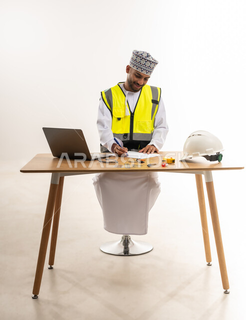 كتابة الملاحظات الخاصة بالمخطط المعماري, دراسة ومتابعة المشاريع الإنشائية, دمج الأجهزة التقنية الحديثة بالأعمال الهندسية, بورتريه لمهندس عربي خليجي عماني يرتدي الدشداشة والكمة مع سترة الحماية يقوم بالتدوين على ورقة بيضاء وبجانبه الحاسوب المحمول, خلفية بيضاء