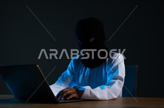 مفهوم الجرائم الالكترونية, القدرة على تهكير الأجهزة, استخدم جهاز الحاسوب لأغراض غير مشروعة, قرصنة وسرقة البيانات, تدمير الحسابات الرقمية, بورتريه مقرب لهاكر عربي خليجي عماني ملثم يرتدي الدشداشة يعمل في مجال الهكر الغير قانوني، خلفية سوداء