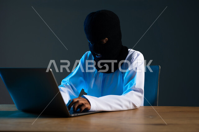 استخدم جهاز الحاسوب لأغراض غير مشروعة، مفهوم الجرائم الالكترونية, القدرة على تهكير الأجهزة, قرصنة وسرقة البيانات, تدمير الحسابات الرقمية, بورتريه مقرب لهاكر عربي خليجي عماني ملثم يرتدي الدشداشة يعمل في مجال الهكر الغير قانوني، خلفية رمادية