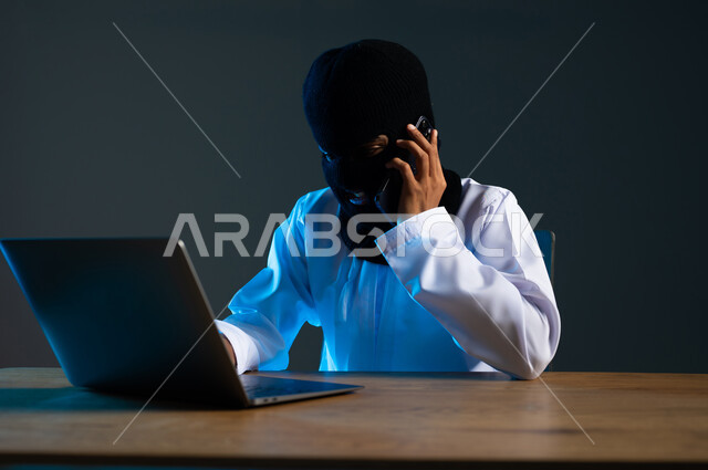التجسس على المكالمات الهاتفية, سرقة وقرصنة البيانات الالكترونية, اختراق الأنظمة وحماية وأمن الأجهزة, استخدام الحاسوب المحمول لأغراض غير شرعية, بورتريه مقرب لهاكر عربي خليجي عماني ملثم يرتدي الدشداشة يمسك بيده الهاتف المحمول، خلفية رمادية