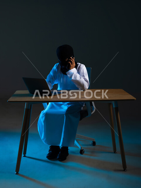 التجسس على المكالمات الهاتفية, سرقة وقرصنة البيانات الالكترونية, اختراق الأنظمة وحماية وأمن الأجهزة, استخدام الحاسوب المحمول لأغراض غير شرعية, بورتريه لهاكر عربي خليجي عماني ملثم يرتدي الدشداشة يمسك بيده الهاتف المحمول، خلفية زرقاء
