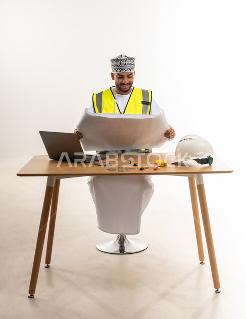 Study and follow-up of construction projects and plans, integration of modern technical devices with engineering works, work in the engineering sector, portrait of an Arab Gulf Omani engineer wearing a dishdasha and a cloak with a protective jacket sitting at the table and holding an architectural plan in his hand, white background