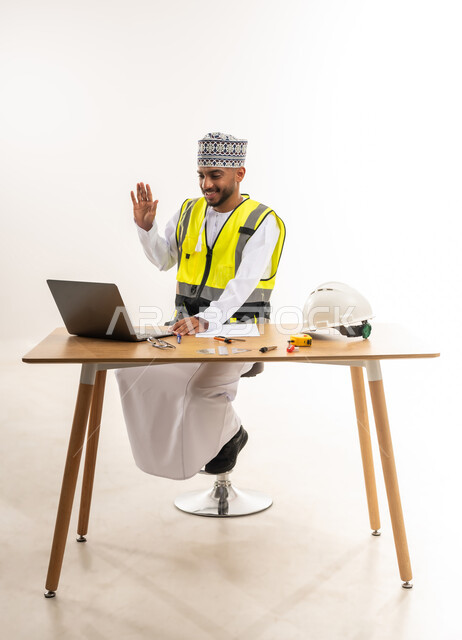 إجراء مقابلة عمل عن بعد, دمج الأجهزة التقنية الحديثة بالأعمال الهندسية, العمل في القطاع الهندسي, بورتريه لمهندس عربي خليجي عماني يرتدي الدشداشة والكمة مع سترة الحماية يجري مكالمة فيديو عبر جهاز الحاسوب المحمول ويرفع يده بتعابير السلام, خلفية بيضاء