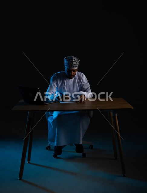 التخطيط للاختراق وسرقة البيانات, اختراق الأنظمة وأمن الأجهزة, خوارزميات لتدمير الحسابات الرقمية, استخدام الحاسوب المحمول لأغراض غير قانونية, بورتريه لهاكر عربي خليجي عماني يرتدي الدشداشة والكمة يقوم بالكتابة على ورقة بيضاء, خلفية سوداء