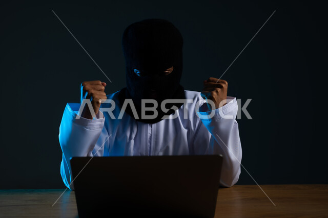 قرصنة وسرقة البيانات, تدمير الحسابات الرقمية, استخدم جهاز الحاسوب لأغراض غير مشروعة، مفهوم الجرائم الالكترونية, القدرة على تهكير الأجهزة, بورتريه مقرب لهاكر عربي خليجي عماني ملثم يرتدي الدشداشة يرفع قبضة يديه بإيماءات الفرح لنجاح عملية الاختراق، خلفية سوداء